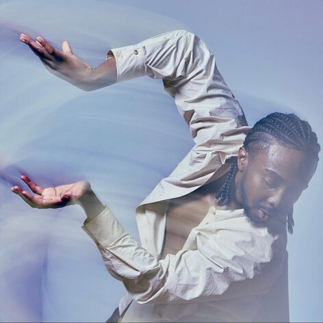 Photo of Stephen Shynes. He holds his arms to the side, hands articulated and elbows bent. He looks towards the ground. He wears an unbuttoned cream top and has his hair in cornrows. Photo by Whitney Browne.
