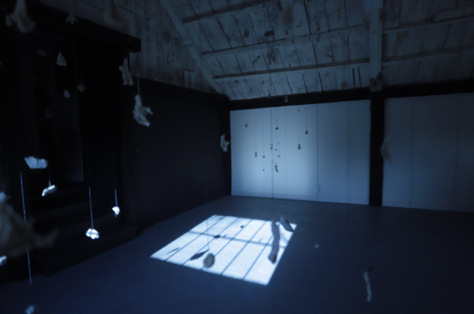Objects attached to strings hang from a white, wooden ceiling in a darkened room. Walking at Night. 2023. Cast dreams into stars into softwood, wood shavings, charcoal and ash. Blue and beige cording. Etched and shaped copper medallions. Photo by Augusta Sparks.