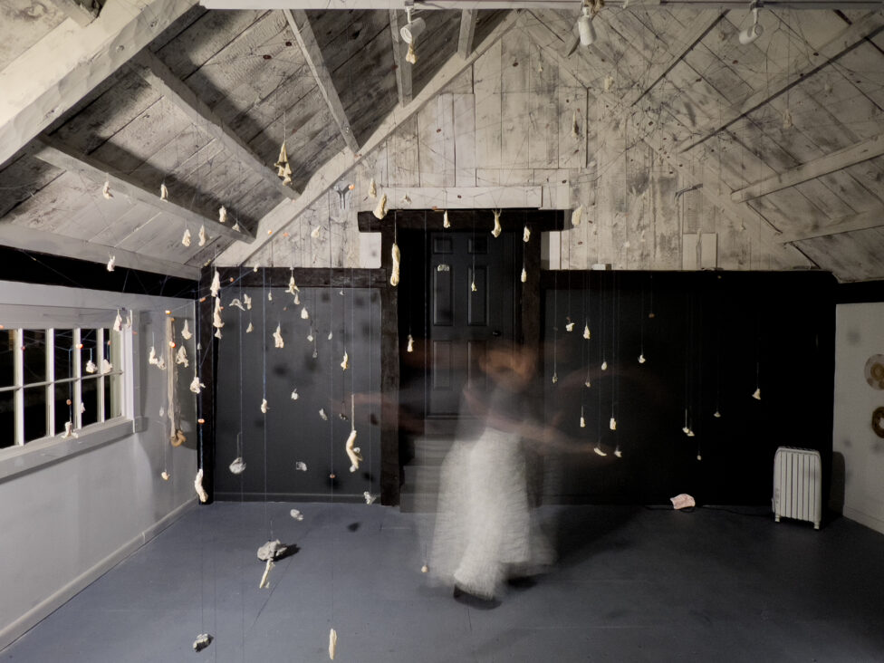 A long exposure photo of Rori Smith moving in a room filled with objects dangling on cording, hanging from the ceiling.