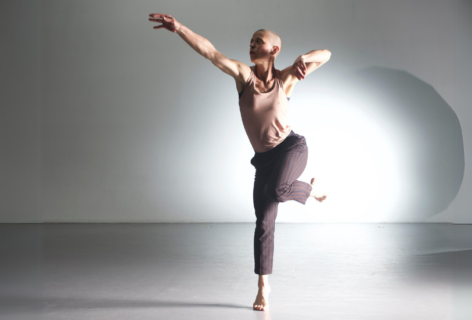 An action shot of Jennifer Nugent in a bright space. A light spotlights behind her as she holds her arm out and balances on one foot. She wears a pink top and grey striped pants.