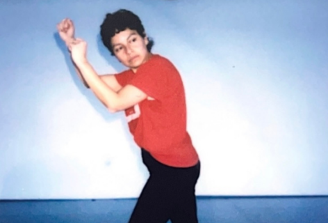 A latina person in a red tee and black leggings wearing white shoes stands in a stepping stance arms in a side motion with fists. She is in a grey dance studio gaze downward and sideways. Photo by Jazzy Romero