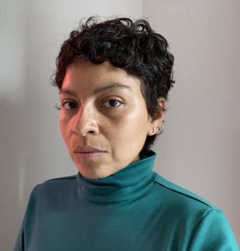 A latina person with black hair and a green turtleneck looks into the camera against a white background. Photo courtesy of Artist.