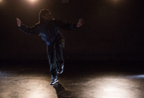Sage Ni'Ja Whitson, a dark skinned Black transgender artist balances on one leg with arms outstretched. They are in a low lit room. Photo by Scott Shaw.