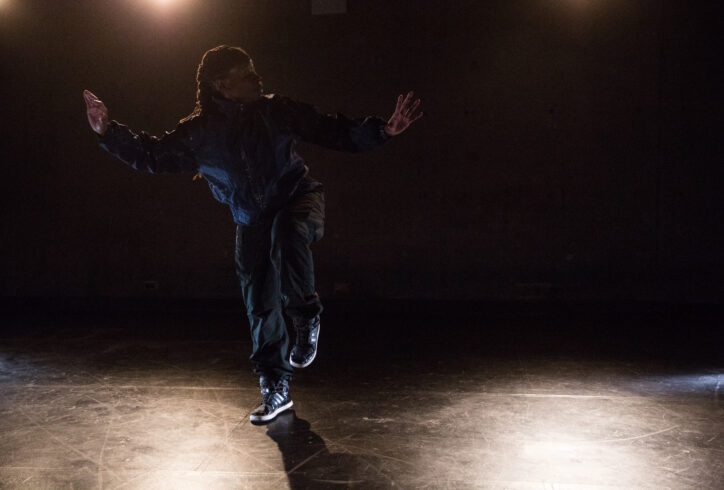 Sage Ni'Ja Whitson, a dark skinned Black transgender artist balances on one leg with arms outstretched. They are in a low lit room. Photo by Scott Shaw.
