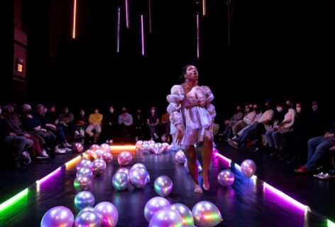 Ogemdi Ude, a dark skinned Black femme, is wearing a puffy purple dress in a room surrounded by colorful balloons and LED floor lights. Her mouth is open with her head tilted up and one leg is raised with the other planted on the floor. Her hands are at her sides. Photo by Maria Baranova.