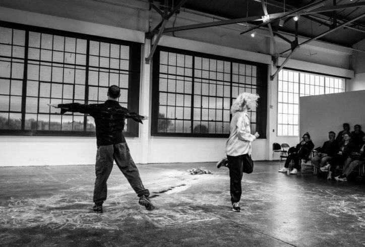 A black and white photo of Sara and Jesse performing at Fort Mason. A warehouse with paneled frosted glass. She wears a white top and black pants. Her hair covers her face while twisting in one direction. Jesse has hie hands out to a T shape and leans in one direction. aA black and white photo of Sara and Jesse performing at Fort Mason. A warehouse with paneled frosted glass. She wears a white top and black pants. Her hair covers her face while twisting in one direction. Jesse has his hands out to a T shape and leans in one direction. Photo by Robbie Sweeney.