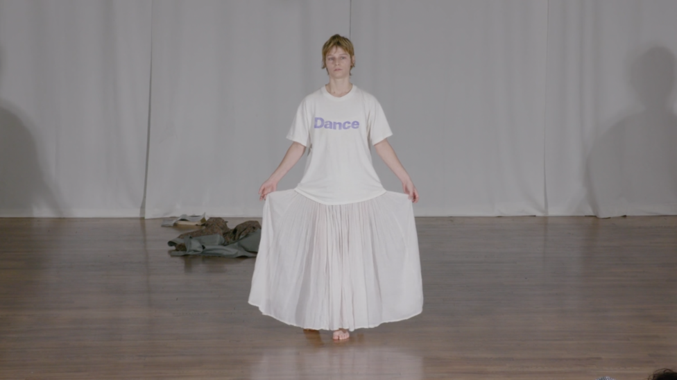 With a dissociated expression, Sonya Gadet-Molansky lifts the two sides of their skirt during a moment in Everything Must Go. They wear a white t-shirt with