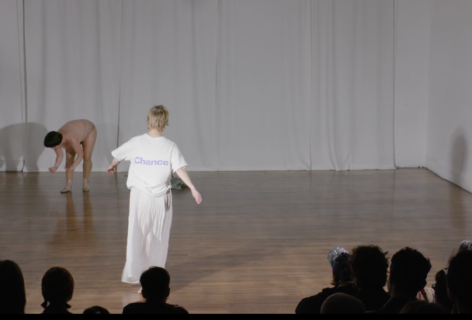 In a white-walled room with brown wooden floor, foregrounded by an audience, two people seem to be doing something. Sonya Gadet-Molansky, downstage, wears a white t-shirt with 
