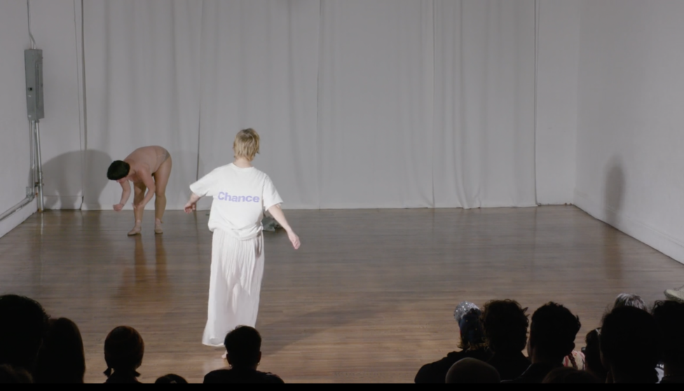 In a white-walled room with brown wooden floor, foregrounded by an audience two people seem to be doing something. Sonya Gadet-Molansky, downstage, wears a white t-shirt with 
