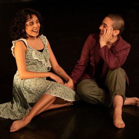 Daniela and Gabby are sitting on the floor towards each other smiling and holding hands. Photo courtesy of the artists.