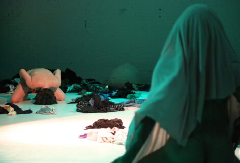 An image from a performance with green lighting and with many costume pieces strewn across the white floor with one performer in the foreground covered by a green lit fabric. In the background diagonally across from is another performer, luciana achugar, kneeling naked on the ground with torso folded forward and crown of head touching the ground with a rounded spine. It looks like prayer or a space for ritual. Photo by Brian Rogers.