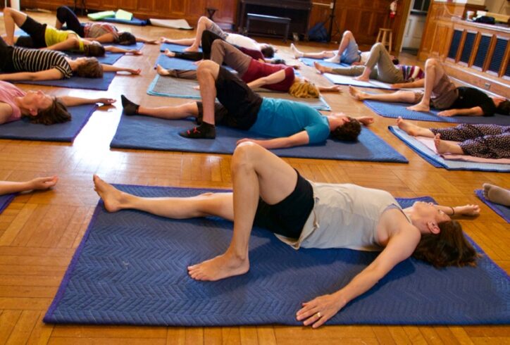 A photo of students practicing movement during a class. They lie on their backs on blue mats. One leg extended downward and the other leg was propped up with their knee bent and the foot pressed into the floor. Photo courtesy of the faculty.