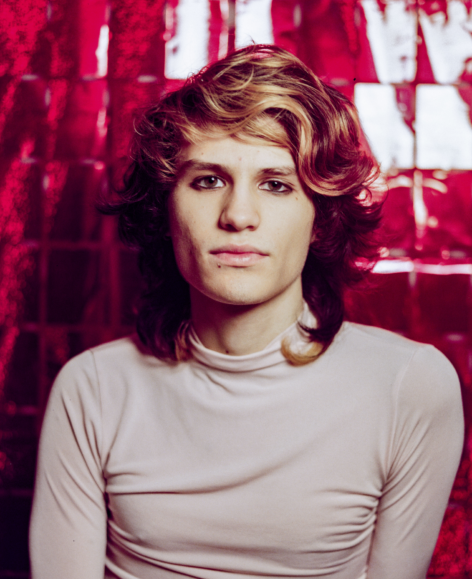 ID: Portrait of a trans-femme identifying individual wearing a pink turtleneck against a red shiny background.