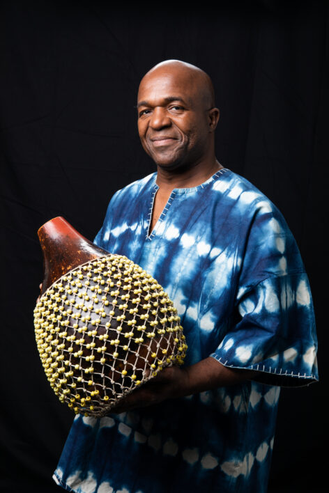 Sherone stands in front of a black backdrop faintly smiling and holding a shekere in his hands.
