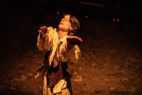 Jo–a white person with brown hair–reaches with both hands toward the camera. Their eyes are closed and there is a yellow cast illuminating their face and clothing. Behind them is a brick wall. Photo courtesy of the artist. 