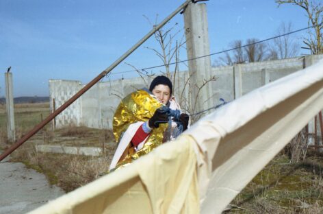 A photo of Chip in a park with a concrete wall behind them. They are wearing a hat and gloves, wrapped in an emergency blanket that they hold in their teeth, while pulling a spool of blue twine between two long metal poles and yellow sheets.