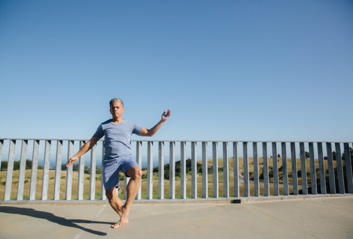 A brown-skinned Filipino man, Gerald wears a blue t-shirt and shorts and dances in front of av blue sky and grey fence. Photo by Crystal Birns