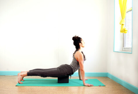 Light skin female is in a plank like position with a block supporting her pubic bone for support. Her chest is extended out and up to a window. Kay Ottinger. Photo Courtesy of artist.