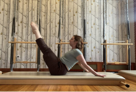 a white woman is balancing on her hips with legs pointed up towards the ceiling, her arms are pressed behind her supporting her. She is seated on a cream mat inside of a Pilates studio. There are wood floors and wallpaper with tree trunks and golden pears on the wall behind her. Photo courtesy of the artist.