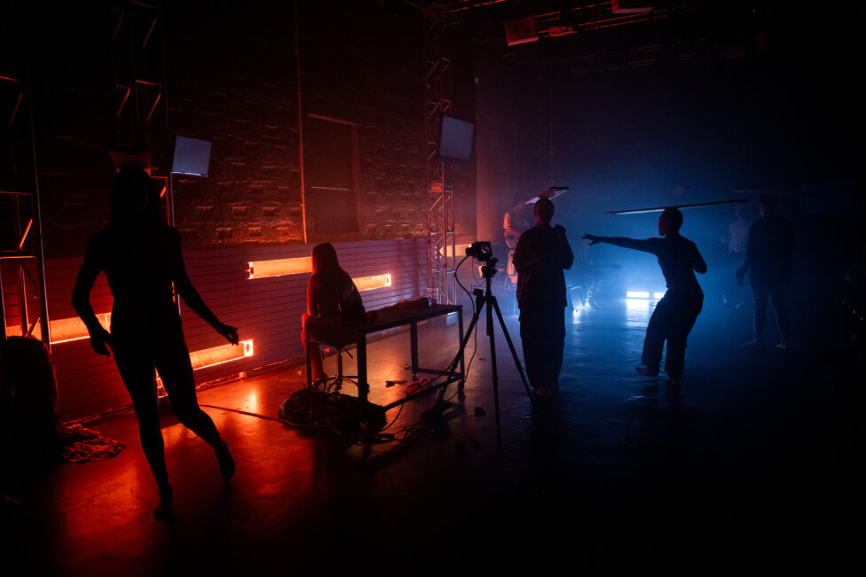On a shadowy stage performers walk balancing planks of wood on their heads. Photo credit to Maria Baranova.