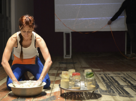 A photo of a light skinned Indigenous female presenting person faces forward and kneels in the left side of the photo. Their head is bent downward with their eyes almost closed. They have dark reddish hair pulled back in a long braid. The braid dangles forward over their rounded shoulders as they lean into a silver bowl. This is moira, their elbows are bent and their hands are in the bowl. The bowl is filled with white colored gooey dough that is drying in large chunks on their hands. They are wearing a white sleeveless tank top, a wide bright orange a pouch at their bent waist and bright blue pants. The bowl sits on a wooden plank floor that has large painted and scuffed shapes in white, black and brown. On moira’s right is a silver tray with holes. It casts a round shadow with dots. On top of the tray are 5 clear glass jars partly filled with gooey liquids and powdery dry grains. The jars have clear gelatin in rounded shapes on top. Each gelatin visible has long green narrow leaves, bright red berries, or a tiny speckled brown egg inside. Behind moira is part of a video projection on a screen. The projection is a close up bird's eye view of a worn stone staircase with a bright orange string slowly moving up the stairs. In front of and to the right of the screen is a live person’s arm stretching in front of the screen and gently holding a bright orange ribbon in their brown hand. The ribbon is attached to the screen and it hangs in 2 big U’s as it dribbles onto the floor. The person appears as a silhouette dressed in black, visible from the shoulders down. Photo by Yuli Dudnyk.