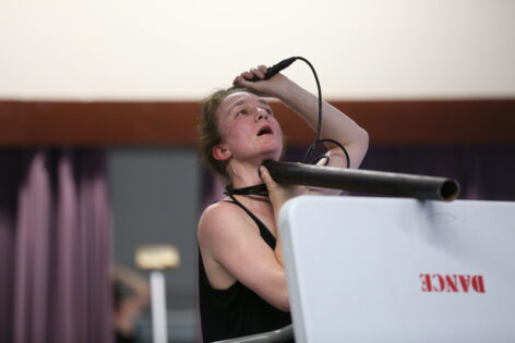 A closeup of Jeanine, holding a metal pipe to her throat with one hand and the other hand holding a mic to her head while she looks up mouth open, standing behind a table with the word