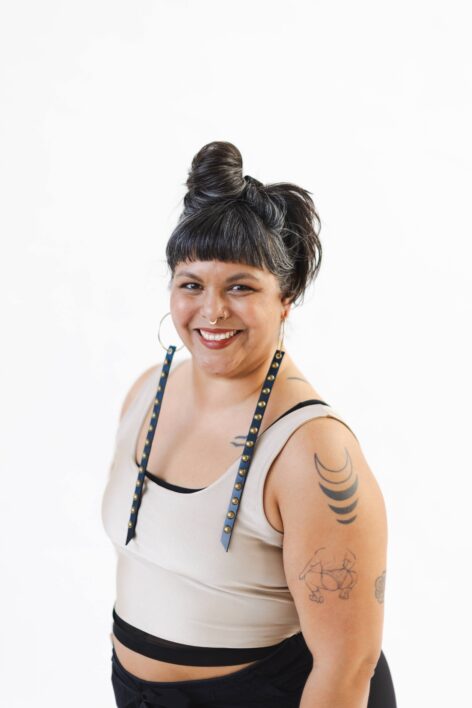 A photo of a cinnamon-colored skinned non-binary femme of Latine descent, born and raised up from the people of the Mapuche, born in Santiago, Chile. This is Taja, they are pictured from their hips up and in a 3 ⁄ 4 pose. Their smiling face is tuned towards us and one slightly tattooed arm hangs along their side. The 3 tattoos are black line drawings that are spaced apart and sit between Taja’s shoulder and elbow. Taja has long, black hair is caressed with stunning silver streaks and short bangs. It is pulled up in a tall bun on the left side of their head. They have black eyebrows, sparkling brown eyes, and a warm beaming smile. Deep red lipstick emphasizes their generous smile. 3 gold rings shimmer in their nose and long black gold studded straps stiffly stretch over Taja’s shoulders. The straps hang from large, thin gold hoops in their ears. They are wearing a white sleeveless cropped tank top layered over a black tank top peaking out at the neck, shoulder and waist. A little bit of their belly peeks out above black pants. The background is white. Photo by Isabel Fajardo.