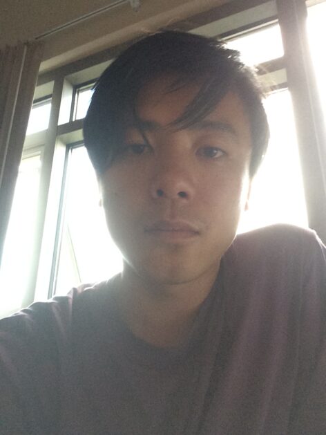 Jian Yi, an East Asian person looks at the camera with short length black hair and purple T-shirt in portrait profile shot as light shines through window behind them. Photo courtesy of the artist.
