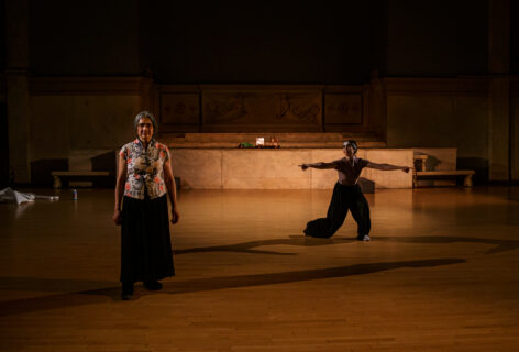 A photo from Melissa Lewis Wong's performance for Movement Research at the Judson Church. One dancers stands facing the audience wearing a floral top. Another stands in a lunge position looking towards the first dancer with their arms spread out away from then with their fingers pointing out. Photo by Rachel Keane.