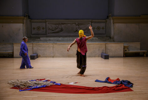 A photo from Jung Woong Kim's work for movement Research at the Judson Church. One dancer in the center wearing a yellow cloth over their face and gesturing upwards and downwards with their arms they wear a red top and brown pants. Another dancer in the background walks toward the first wearing a purple outfit. In the foreground a large red cloth is spread on the ground along with strips of blue fabric and beige material. Photo by Rachel Keane