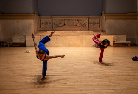 Daniela Garcia-Arce and Gabby Gubitosi perform for Movement Research at the Judson Church. They balance on one leg torso leaning forward and counter balanced by their other leg swinging upwards with a bent knee. Photo by Rachel Keane.