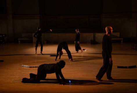 Luis A. Lara Malvacias' work for Movement Research at the Judson Church. Five dancers all in black clothing move around the space. Some walk on is on their hands and knees and another bends over pushing this his upward. Photo by Rachel Keane.