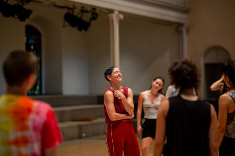 Jennifer a white woman with a black bandana wearing red pants and a red tank top is smiling large / laughing and gathered with a group of MELT participants at St. Marks Church in the Bowery. Photo by Rachel Keane.