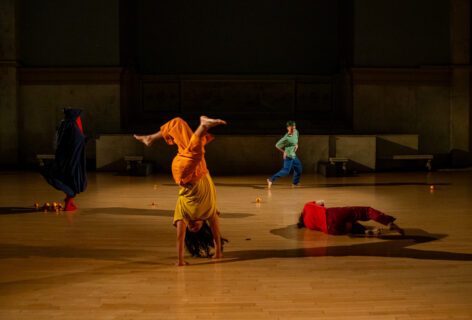 Photo from Paris Cullen's performance at Movement Research at the Judson Church. Dancers move around the space each wearing bold colorful costumes. One person runs, another moves on the ground, another stands covered by a dark blue fabric and a person supports their body in a headstand. Photo by Rachel Keane.