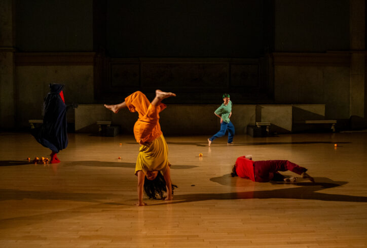 Photo from Paris Cullen's performance at Movement Research at the Judson Church. Dancers move around the space each wearing bold colorful costumes. One person runs, another moves on the ground, another stands covered by a dark blue fabric and a person supports their body in a headstand. Photo by Rachel Keane.