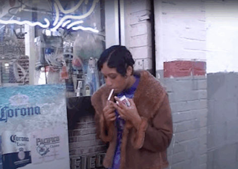 Benin in a fur coat lights a cigarette in front of a store window. Photo courtesy of artist.