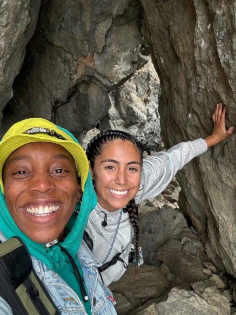 Bria Barfield and Brianna Torres. (left to right) Two women smile at the camera, Brianna's hand placed on the rock. They are standing in between a coastal rock formation in Marin County, CA. Photo courtesy of the artists.
