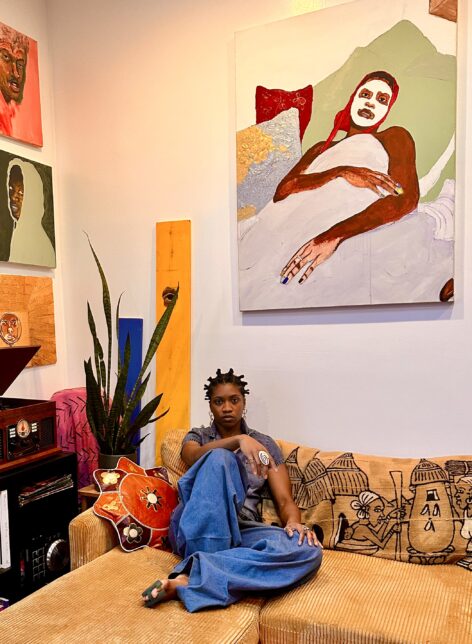 Dorchel Haqq sits in a denim outfit on a mustard couch. Above them paintings hang, and beside them are pillows with patterns. They look toward the camera with a hand on their knee and arm on the couch. Photo by Hakeem Olayinka.