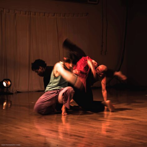 An ensemble of people dancing in contact together layered on top of one another, with a blurry motion in a shadowy theatre. Photo courtesy of the artists.
