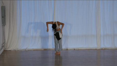 Benin stands against a white curtain wearing a cropped white tank top and denim pants with cuttings that reveal black shorts underneath. Her feet are together and her upper body is draped over. Both elbows are lifted as her hands point towards the floor. Photo by Kayhl Cooper.