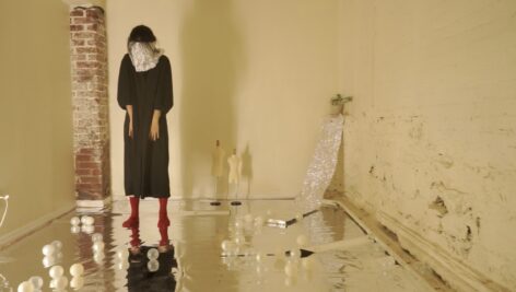 Zhangxinan Wu with aluminum foil covering her neck, dropping her head into the neck holder. She is standing on reflective foil surface with balls in different shades of white rolling on the floor. Photo by Bella Thorpe-Woods.