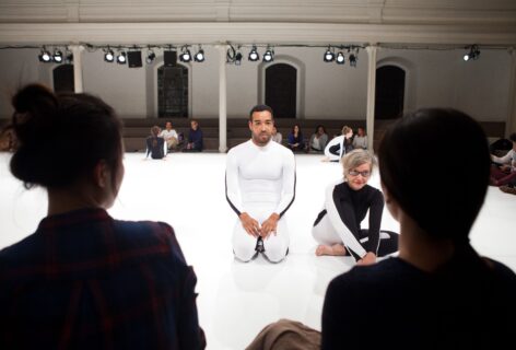 2 performers sit on a white marly floor in all white and all black unitards. The silhouette of the back 2 people's heads in the foreground and groups of people in the background behind the seated dancers. Photo by Miana Jun.