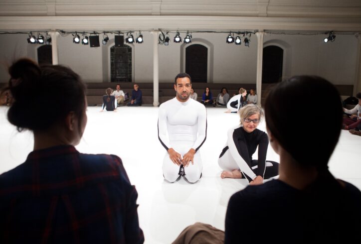 2 performers sit on a white marly floor in all white and all black unitards. The silhouette of the back 2 people's heads in the foreground and groups of people in the background behind the seated dancers. Photo by Miana Jun.