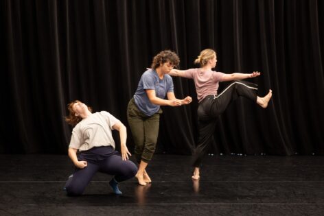 Three performers are captured mid-motion. One dancer kneels and tosses her head back. The second dancer balances with her legs closely crossed, pinching her hands and looking toward the floor. The third dancer faces the back with both arms extended at shoulder height, raising a bent leg to the side with a flexed foot.