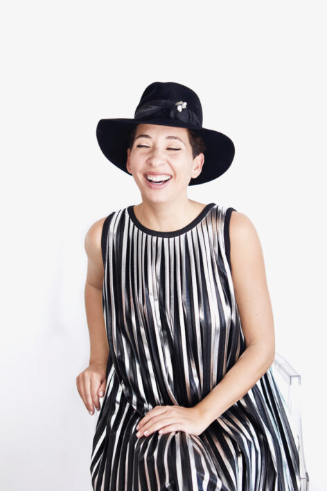 Portrait of Shirine in a sleeveless striped blouse and black hat mid laughter. Their eyes are slightly closed while they sit with their hand on their knee crossed over the other in front of a white background. Photo credit: Eva Sakellarides.
