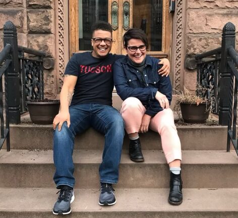 Two people, one with their arm around the other and both wearing glasses, are sitting on a stoop in front of a Brownstone in Brooklyn looking at the camera and smiling. Marci, a nonbinary, brown skin person with short cropped brown hair, longer on top and shaved on the sides is wearing a black T-shirt with the word Tucson spelled out in red all cap letters across their chest, a pair of faded blue jeans and black and grey running shoes. Diana is wearing a black jean jacket over a grey turtleneck sweater, pink jeans and a pair of ankle high black leather boots with white socks peeking out. Photo credit: Micheal O'Neill.