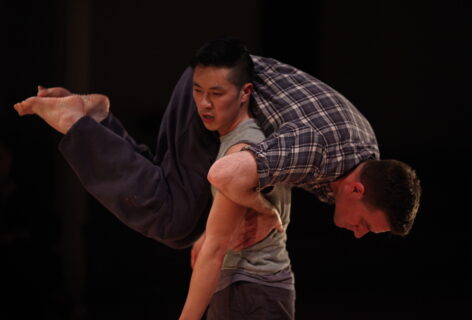 A photo of two dancers. One dancer carries the other over their shoulder. The second dancer places one hand on his partner's waist for support, their legs are bent. Photo by Paula Court .