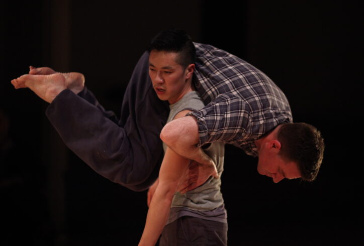 A photo of two dancers. One dancer carries the other over their shoulder. The second dancer places one hand on his partner's waist for support, their legs are bent. Photo by Paula Court .