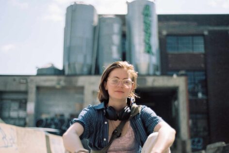 A white person with mid length ginger hair sitting outside in front of a blurry factory. They're wearing a blue shirt and have headphones around their neck. Photo credit to Ixc Wylde.
