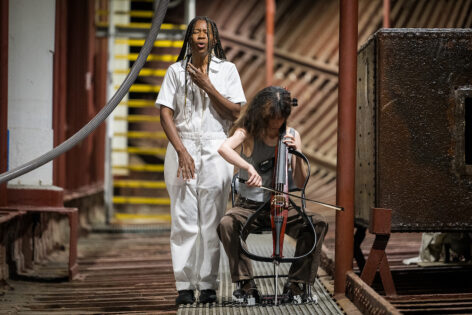 A brown skinned performer with long brown braids singing in a white jumpsuit with an open left hand gesture framing the face and the other hand reaching down towards the ground. They are standing next to light skinned person with long hair and bangs, head down in full concentration playing the electric cello; both performers are in the cargo hold of a large ship with brick red beams on either side and an old steel furnace on the left and behind them is a yellow staircase. Photo by Cherylynn Tsushima.